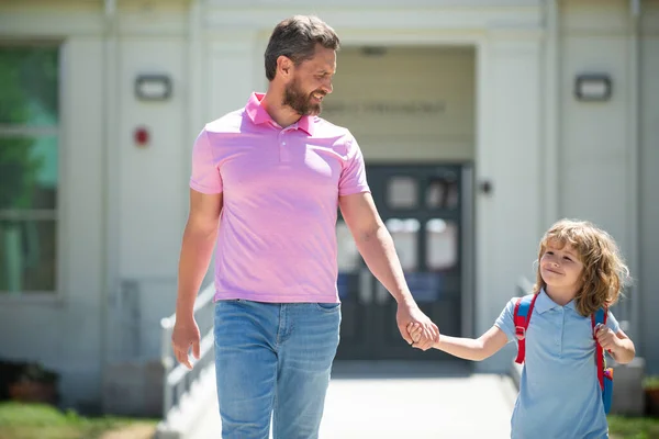 Parent and pupil of primary school go hand in hand. Teacher in t-shirt and cute schoolboy with backpack near school park. — Stock Photo, Image