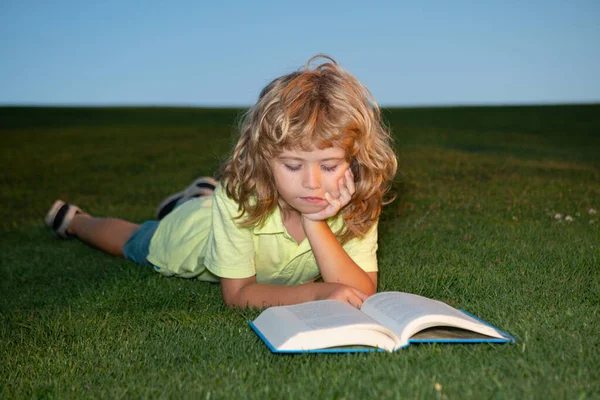 Elinde kitap olan okul çocuğu dışarıda dinleniyor. Çocuk bahar parkı hakkında bir kitap okuyor. Bahçedeki çimlerin üzerinde dinleniyor. Açık hava eğitimi. — Stok fotoğraf