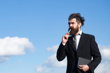 Gökyüzü olan bir iş adamı. İşadamı kalemle ilgili bir şey düşünüyor. İş hayalleri. Tişörtlü ve kravatlı düşünceli genç adam elinde not defteri ve kalemle.