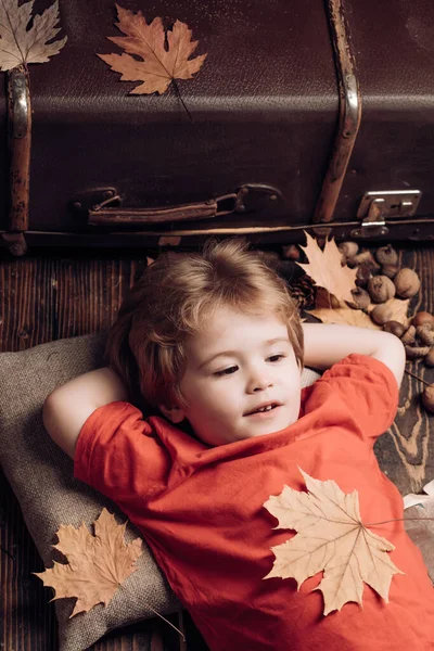 Blond liten pojke vilar med löv på magen ligger på trägolv på höstlöv. Liten pojke ligger på en varm filt drömmar om varm höst. Ungen leker på hösten. — Stockfoto