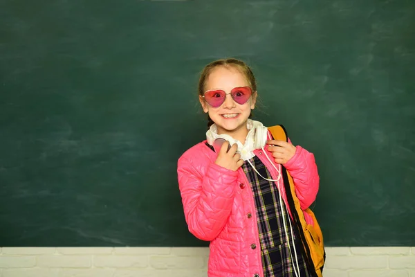 Lächelndes kleines Schulmädchen mit Rucksack hält Bücher an die Tafel. Lebensstil in der Kindheit. Bildung in der Schule. — Stockfoto