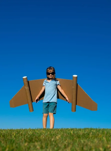 青い空に紙の翼を持つ子供の男の子。春の緑の野外で楽しいおもちゃのジェットパックを持つ子供。自由気楽で子供の想像力夢のコンセプト. — ストック写真