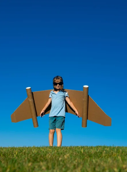 Niño con alas de papel contra el cielo azul. Niño con juguete jetpack divertirse en primavera campo verde al aire libre. Libertad despreocupado y niños imaginación sueño concepto. — Foto de Stock