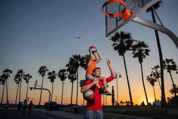Αστείο ομαδικό μπάσκετ, αγόρι με προπονητή. Εκπαιδευτής κρατώντας το παιδί στους ώμους, βοηθώντας τον να σκοράρει ένα καλάθι σε μια παιδική χαρά. Εκπαίδευση σε σχολή μπάσκετ. — Φωτογραφία Αρχείου