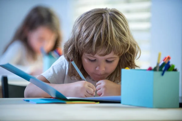 Школьник что-то пишет в блокноте и сидит за столом в классе. Возвращайся в школу. Счастливый улыбающийся щенок рисует на столе. Ребенок из начальной школы в классе. — стоковое фото