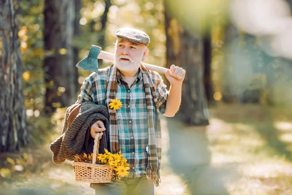 Bearded man relax in forest. Smiling old forester with axe and flowers. Happy man with beard and mustache hold axe. Elderly male walk in forest. Bearded man in hat on a background of trees. — Stock Photo, Image