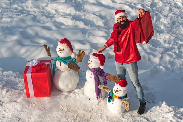 Hombre excitado con muñeco de nieve divertido en sombrero elegante y bufanda en el campo cubierto de nieve. Feliz invierno muñeco de nieve de la familia con regalo. Madre de la nieve-mujer, el padre de la nieve-hombre y el niño desea feliz Navidad y Feliz Año Nuevo. — Foto de Stock