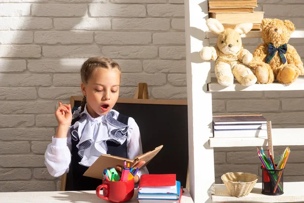 Poważnie zabawna uczennica czyta książki w klasie w szkole. Dzieciak uczy się w szkole. Uczennica odrabiała lekcje w klasie. Edukacja dla dzieci. Niesamowita twarz dzieci. — Zdjęcie stockowe