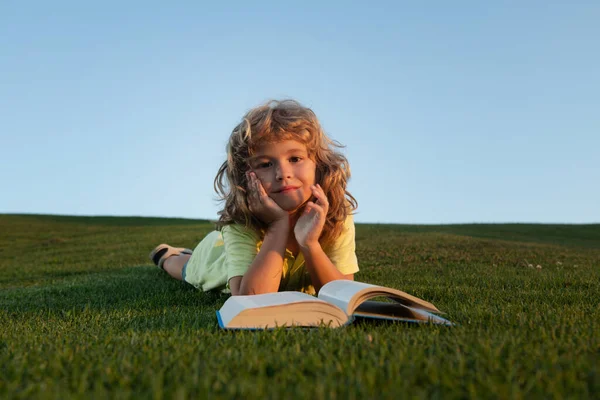 Niño leyendo libro que pone en la hierba sobre la hierba y el fondo del cielo con espacio de copia. El colegial con el libro que descansa al aire libre. —  Fotos de Stock