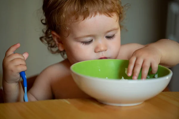 Ritratto di bambino caucasico carino con cucchiaio. Bambino affamato disordinato con piatto dopo aver mangiato purea. — Foto Stock