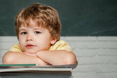 Okul malzemeleriyle masasının yanında duran mutlu yüzlü bir öğrenci. Çocuk arkaplanda bir tahtayla cevap vermeye hazır. Öğretmenler Günü.