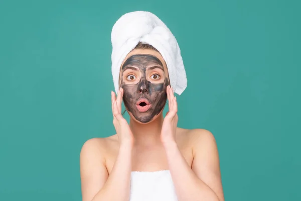 Överraskad kvinna med lera ansiktsmask, ansikte lera mask spa. Vacker kvinna med kosmetisk lera ansiktsbehandling, spa hälsa koncept. Skönhetsbehandling av huden. Handduk på huvudet. Medicinskt minerallera. — Stockfoto