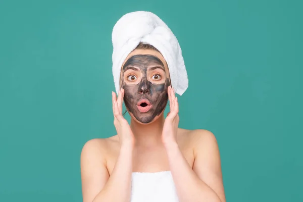 Wanita yang terkejut dengan masker wajah lumpur, wajah masker tanah liat spa. Wanita cantik dengan prosedur wajah lumpur kosmetik, konsep kesehatan spa. Perawatan kecantikan perawatan kulit. Handuk di kepala. Lumpur mineral medis. — Stok Foto