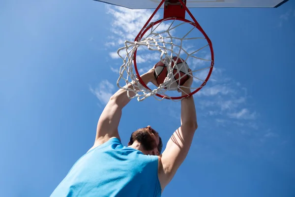 Basketspelare. Sport och basket. En ung man hoppar och kastar en boll i korgen. Blå himmel och domstol i bakgrunden. — Stockfoto