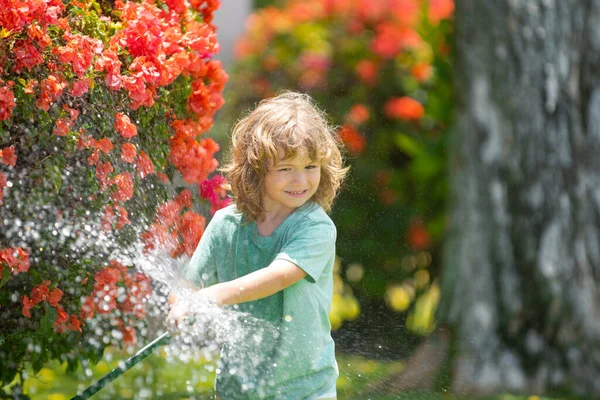 Kid playing in garden, pours from the hose, makes a rain. Happy childhood concept. Child pouring water on the trees. Kid care for the plants in backyard. — Stock Photo, Image