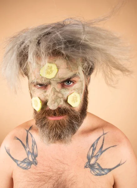 Funny man with clay mask and cucumber slices on face. Male spa, dermatology, wellness and facial treatment concept. Man having cosmetic moisturizing mask, isolated beige background. Male face care. — Stock Photo, Image