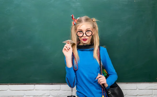 Portret van een tienerstudente. Leuke tienerstudente. Aantrekkelijk jong meisje met rugzak en papieren vliegtuig, staande tegen schoolbord. — Stockfoto