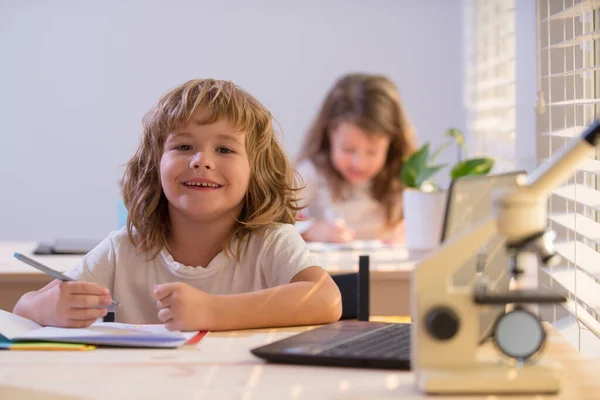 Niño de la escuela sentado en la mesa, escribiendo deberes o preparándose para el examen. Feliz niño sonriente estudio. Poco aprendizaje de los estudiantes. — Foto de Stock