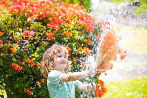 Lindo niño regando flores en el jardín en el día de verano. Niño agricultor con manguera de jardín en planta de plantación. Manguera. Divertido niño regando plantas en el jardín del patio. —  Fotos de Stock