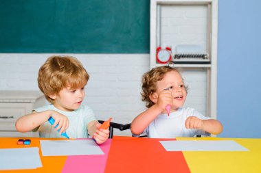 Neşeli, gülümseyen küçük bir oğlan ve küçük tatlı bir kız tahtaya karşı eğleniyor, okul kavramı. Okula ilk gelişim. Okul malzemeleriyle masasının yanında gülen surat ifadesiyle küçük bir çocuk..