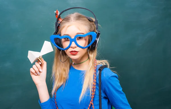 Portret van een tienerstudente. School tiener meisje met rugzak en papier vliegtuig, staande tegen schoolbord. — Stockfoto