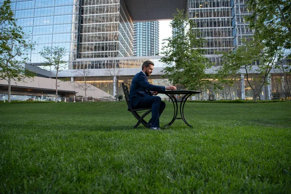 Businessman on large grass field near modern office building. Businessman with laptop in park of megapolice.