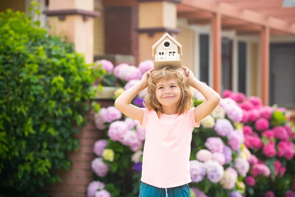 Bienes raíces, vivienda y concepto familiar. Divertido niño sosteniendo juguete casa de madera cerca de nuevo hogar. — Foto de Stock