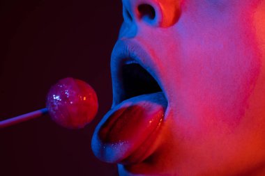 Ağzında lolipop var, yakın plan. Ağzında lolipop olan güzel bir kız. Dili parlak kırmızı kadın dudakları. Ağız yalamak neon ışıklarda çene emmek.