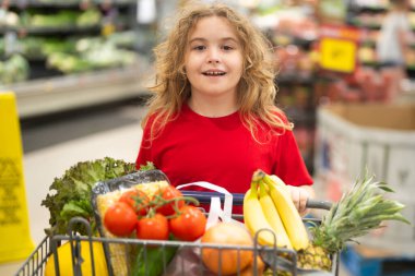Süpermarkette el arabasıyla çocuk alışverişi. Çocuk markette taze sebze seçiyor. Süpermarkette yiyeceği olan bir çocuk. Süpermarkette ürün seçen bir çocuk. Süpermarkette taze yiyeceği olan çocuk