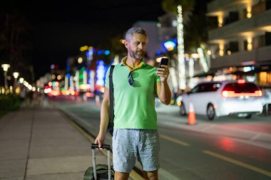 Gece Miami sokaklarında telefon kullanan bir gezgin. Erkek turist yaz macerası sırasında telefonla arıyor. Telefonda sıradan bir turist konuşması. Gezgin telefonunu kontrol ediyor. Tropik bir şehirde bavullu bir adam.