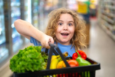 Markette alışveriş arabası olan bir çocuk. Çocuk taze sebze seçiyor. Yakışıklı çocuk sağlıklı yiyecek için alışveriş yapıyor. Süpermarketteki çocuk. Meyve pazarındaki çocuk. Çocuk marketten sebze alıyor.