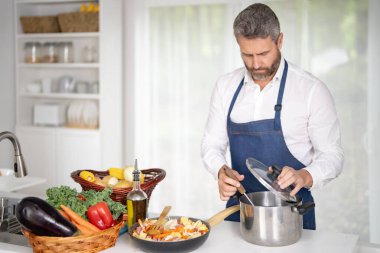 Taze sebze pişiren bir şef. Ev yapımı yemek pişiren bir adam. Mutfakta sağlıklı bir kahvaltı için sebze pişiren bir aşçı. Üniformalı bir adam beyaz bir evde sebze pişiriyor.