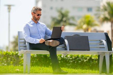 Kendine güvenen işadamı bankta dizüstü bilgisayarı ve parktaki telefonuyla oturuyor. Başarılı bir iş adamı. Olgun iş adamı tatilin tadını çıkarıyor. İş adamı parktaki bankta mola veriyor. Yaz işleri.