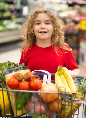 Süpermarkette el arabasıyla çocuk alışverişi. Çocuk markette taze sebze seçiyor. Süpermarkette yiyeceği olan bir çocuk. Süpermarkette ürün seçen bir çocuk. Süpermarkette taze yiyeceği olan çocuk