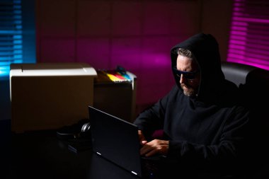 Bilgisayar korsanları siber saldırı sırasında bir gece ofisindeki dizüstü bilgisayarda yazarlar. Siber suçlu ve çalıntı finansal veri konsepti. Dijital para hırsızı. Siber saldırı. Ofiste laptop kullanan kapüşonlu bir bilgisayar korsanı.