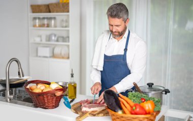 Adam mutfakta sebze doğruyor. Önlüklü aşçı modern mutfakta sağlıklı yemek hazırlar. Mutfakta sebzeli bir aşçı. 40 'lı yaşlar ev mutfağında sebzelerle kahvaltı hazırlar.