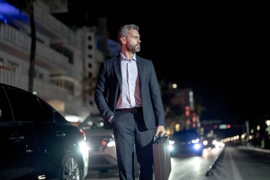 Gece vakti takım elbiseli bir iş adamı şehirde yürüyor. Takım elbiseli zengin bir adam. Lüks arabanın yanında bir iş adamı. Olgun bir iş adamı gece şehir ışıklarında yürür. İş stili. Zengin iş adamı