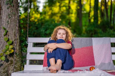 Kaygısız çocuk arka bahçede salıncakta dinleniyor. Çocuk salıncakta rahat ve neşeli oturuyor. Küçük çocuk dışarıda eğlenmenin ve eğlencenin tadını çıkarıyor. Çocuk sakince sallanıyor yeşil bahçede