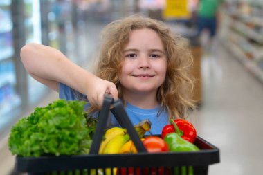 Marketteki çocuk. Sebze dolu alışveriş arabası olan çocuk. Çocuk sebze seçiyor. Süpermarkette çocuk alışverişi. Çocuklar için market eğlencesi. Bakkaldaki çocuğun yüzü. Çocuk hipermarkette