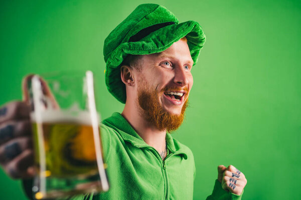 Лепрекон с радостью держит кружку пива на день Святого Патрика. Зеленый костюм для празднования Святого Патрика. Человек наслаждается праздничным напитком в праздник Святого Патрика