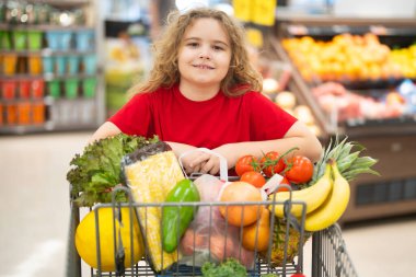 Süpermarkette el arabasıyla çocuk alışverişi. Çocuk markette taze sebze seçiyor. Yiyecek alışverişi yapan çocuk. Yerel bir dükkanda sağlıklı ürünler seçen bir çocuk. Hipermarkette taze malları olan çocuk