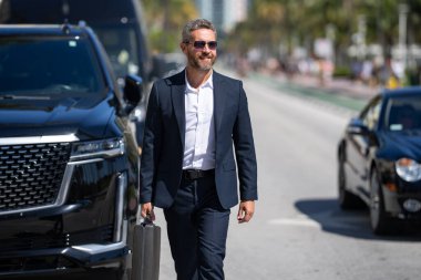 Lüks bir arabanın yanında zengin bir iş adamı. Takım elbiseli zengin bir iş adamı. Şehirdeki başarılı zengin iş adamı. Kendine güvenen bir iş adamı. Başarı işi ve zenginlik. İş adamı sokakta yürüyor