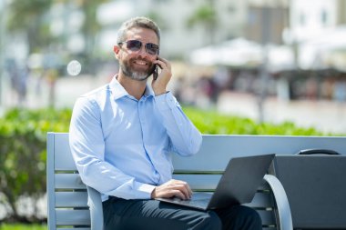 Kendine güvenen bir iş adamı bankta dizüstü bilgisayarı ve parktaki telefonuyla oturuyor. İspanyol iş adamı. Olgun iş adamı tatilin tadını çıkarıyor. İş adamı parktaki bankta mola veriyor. Yaz işleri.