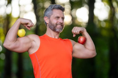 Güçlü bir adam pazılarını elmalarla esnetir. Olgun sporcu, parkta egzersiz yaparken pazılarını gösteriyor. Orta yaşlı bir sporcu pazıları eğitir. Egzersiz sonrası erkek pazıları