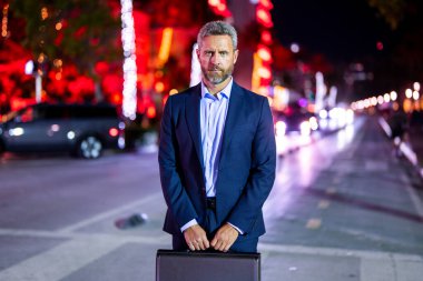 Gece vakti evrak çantalı bir iş adamı. Şehirde takım elbiseli zengin bir adam. Lüks arabanın yanında bir iş adamı. Şehir ışıklarının yanında yürüyen bir iş adamı. İş stili. Lüks ortamda zengin bir işadamı