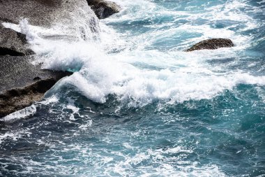 Okyanus dalgaları kayalık kıyı şeridine çarpıyor. Deniz dalgası sıçrıyor. Mavi turkuaz su dalgası deniz kenarındaki kayalar. Köpüklü ve dalgalı deniz manzarası. Dalgalar dokulu kaya yüzeyinde kırılıyor