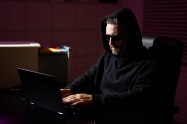 Karanlık bir ofiste dizüstü bilgisayarı olan bir bilgisayar korsanı. Gecenin geç saatlerinde kapüşonlu bir bilgisayar korsanı verileri hackliyor. Siber suçlu ve bilgisayar korsanı çalıntı dijital bilgi konsepti. Hacker gece ofisinde dizüstü bilgisayar kullanıyor.