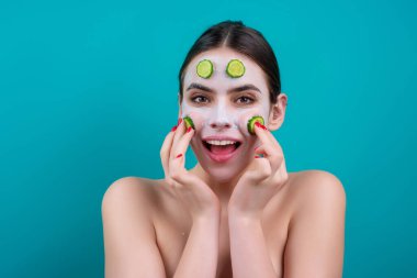 Woman with facial mask holds slices of cucumber, over blue background. Beautiful young woman with facial mask with cucumber. Skin care and treatment, spa, natural beauty and cosmetology