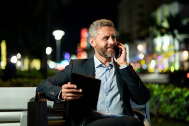 İş adamı telefonda konuşuyor ve tablet kullanıyor. Tabletli bir iş adamı. Tabletli ve telefonlu bir iş adamı. Tablet kullanan bir işadamı, bankta oturur. Gece işi.