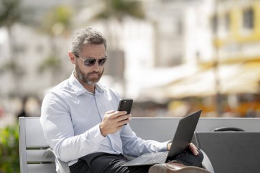 Kendine güvenen bir iş adamı bankta dizüstü bilgisayarı ve parktaki telefonuyla oturuyor. Zengin iş adamı. Olgun iş adamı tatilin tadını çıkarıyor. İş adamı parktaki bankta mola veriyor. Yaz işleri.